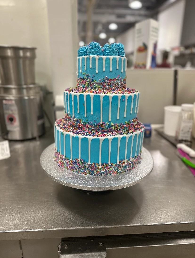 Three-tier blue cake with white drips, rainbow sprinkles, and blue buttercream rosettes on top