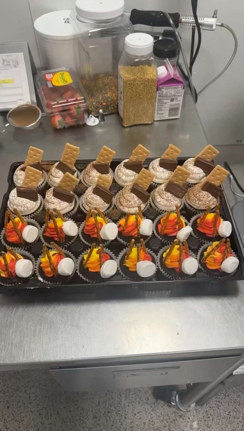 Tray of campfire and s'mores cupcakes topped with graham crackers, chocolate squares, marshmallows, and pretzel sticks