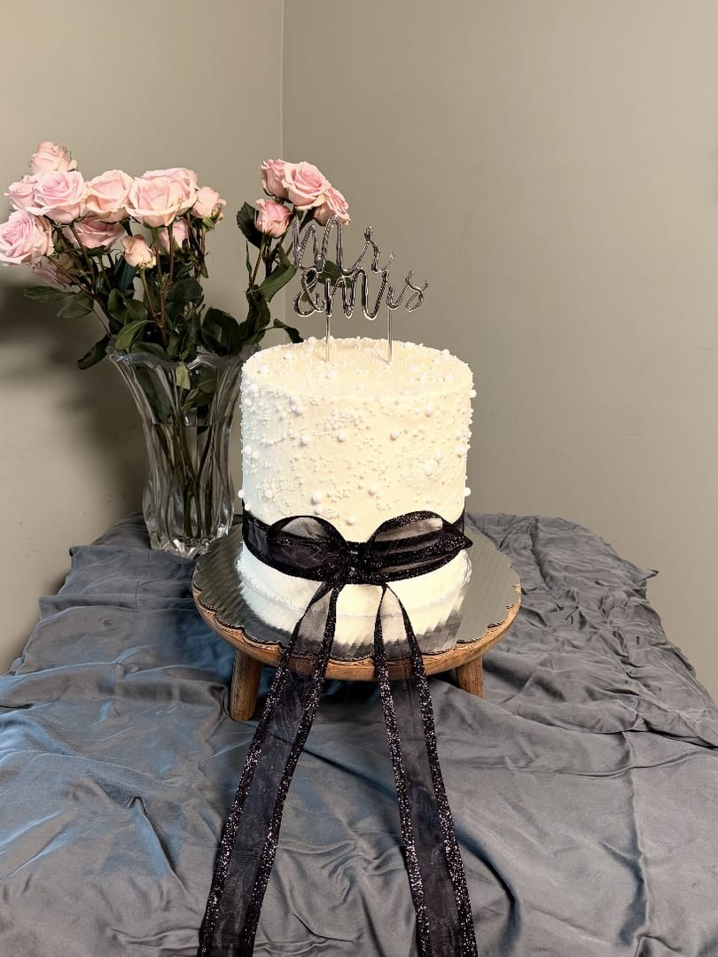 Tall white textured cake tied with a black ribbon beside a vase of pink roses