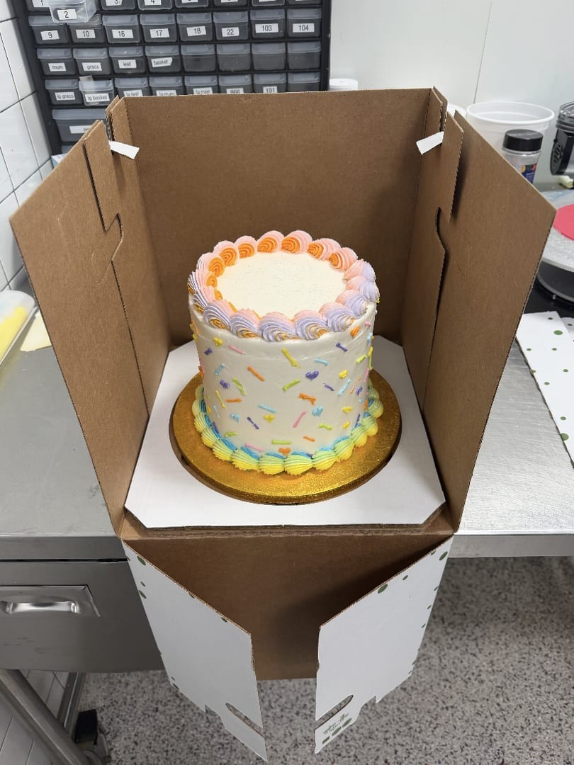 Small round cake with a pastel scalloped border and sprinkle pattern, in a cake box